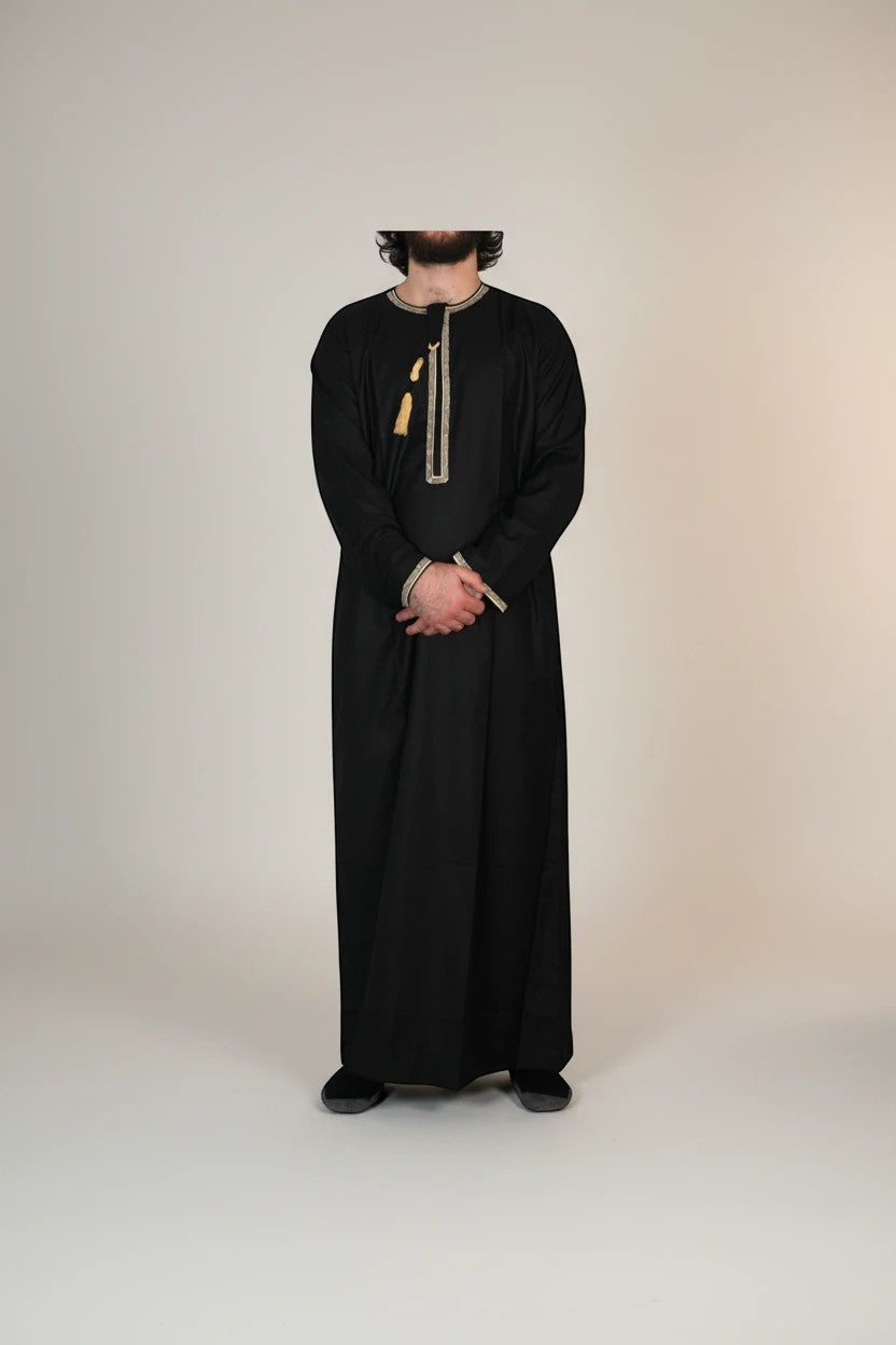 Khayr Thobes black traditional thobe with gold and black embroidered neckline and tassels. Long sleeves, flowing silhouette.