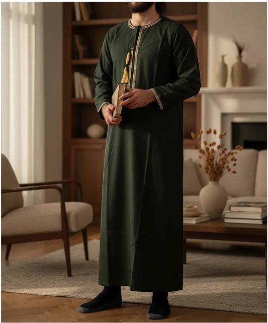 A man wearing a dark forest green long-sleeve thobe with gold embroidery and a gold zipper detail, standing in a warm, modern living room.