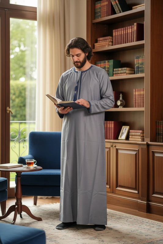 Khayr Thobes grey long sleeve thobe with navy trim and tassel detail, worn by a man reading a book.